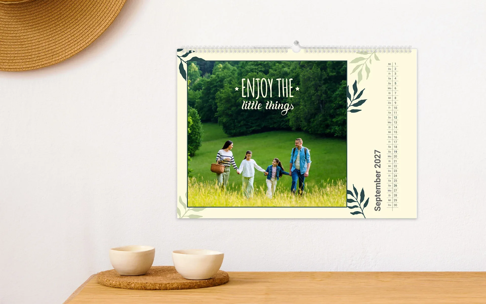 HappyFotokalender ca. A4 quer – aufgehängt Ein aufgehängter HappyFotokalender ca. A4 quer mit dem Text Enjoy the little things zeigt eine Familie auf einer Wiese und Blätter Cliparts.