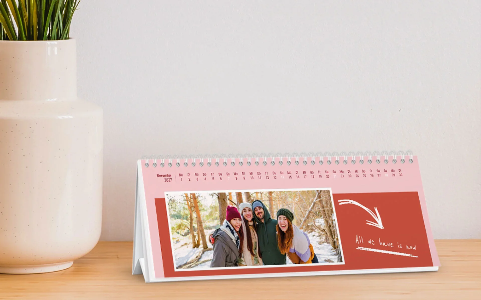 Ein aufgestellter Tischkalender mit dem Text All we have is now zeigt 4 Freunde im Wald vor einem rosa Hintergrund und einem roten rechteckigen Clipart.